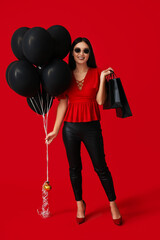 Beautiful young Asian woman with shopping bags and balloons on red background. Black Friday sale