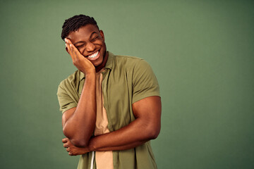 Positive reaction. Afro american guy touching cheek with hand while pleasantly amazed with something in studio. Happy young man with closed eyes and toothy smile expressing joy over green background.