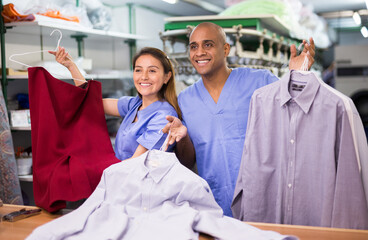 Man and woman dry cleaners operators posing together showing clean clothes on hangers