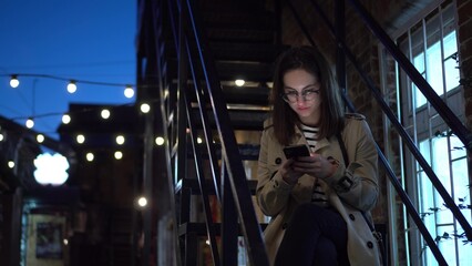 A young woman sits on a fire escape and texts on a smartphone. A girl with glasses on a narrow street late at night with a phone in her hands.