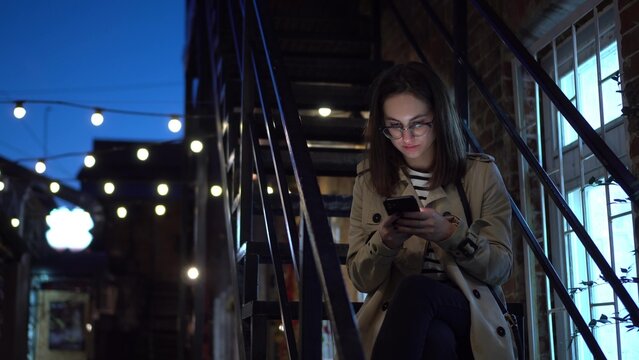 A Young Woman Sits On A Fire Escape And Texts On A Smartphone. A Girl With Glasses On A Narrow Street Late At Night With A Phone In Her Hands.