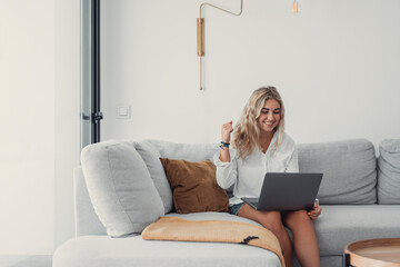 One young happy woman winning and having fun celebrating on couch using laptop pc at home. Euphoric...