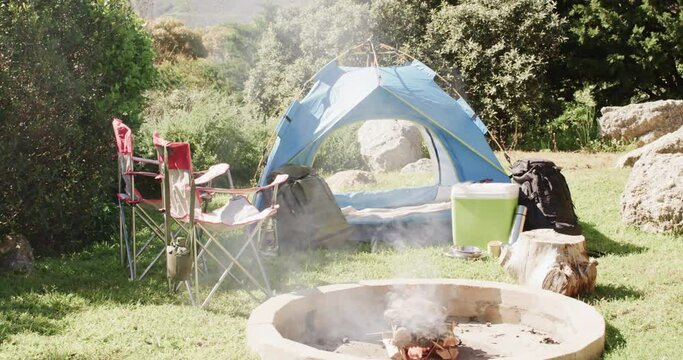 Smoking firepit, tent, chairs and camping equipment in sunny countryside, copy space, slow motion