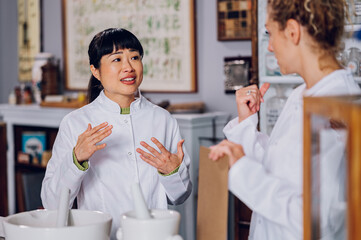 A Japanese chemist is standing with her assistant in the pharmacy and teaching her the cure-making process.