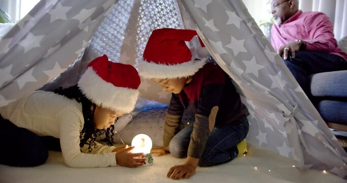 African american brother and sister in christmas hats using snow globe in blanket tent, slow motion