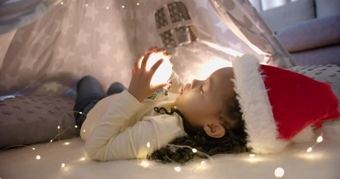 Happy african american girl in christmas hat holding glowing snow globe in blanket tent, slow motion