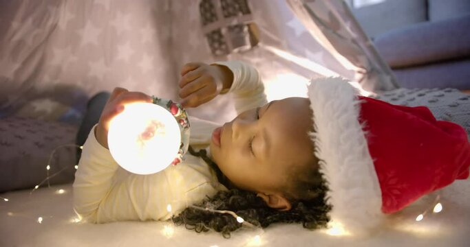 Happy african american girl in christmas hat holding glowing snow globe in blanket tent, slow motion