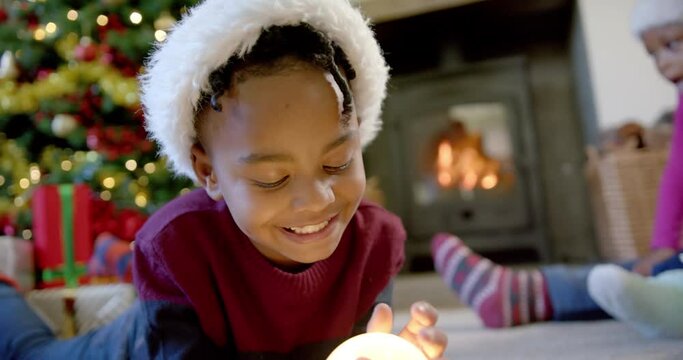 Happy african american son in christmas hat holding snow globe, family in background, slow motion