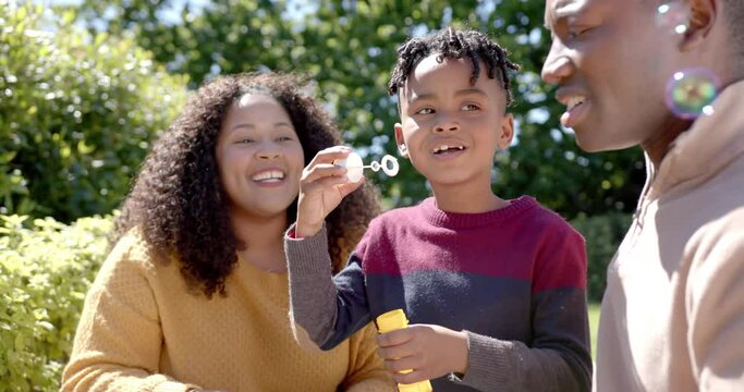 Happy african american mother and father with son blowing bubbles in sunny garden
