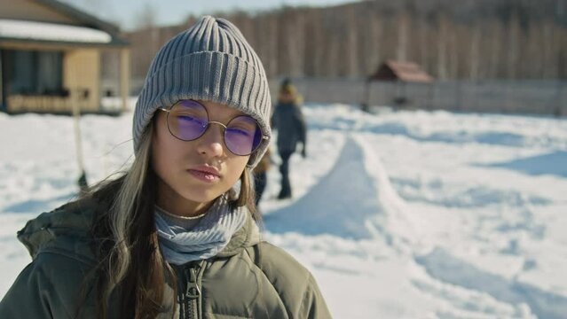 Medium portrait shot of teenage Caucasian girl in woolly hat, glasses and coat standing outdoors in yard on sunny winter day and looking at camera, and friends making snow castle in background