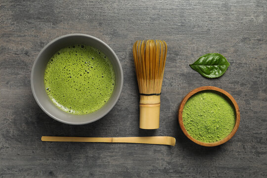Cup of fresh matcha tea, bamboo whisk, spoon and green powder on dark grey table, flat lay