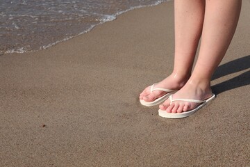 Fototapeta na wymiar Woman in stylish white flip flops on sandy beach near sea, closeup. Space for text