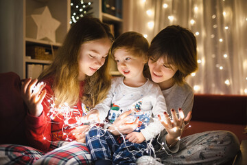 Two big sisters and their toddler brother playing with Christmas lights in a cozy living room on...