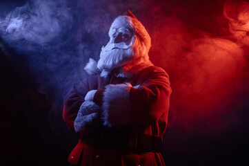 Santa Claus stands with his arms crossed on his chest in blue red neon light and smoke. 