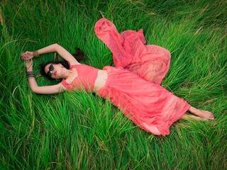 Attractive young brunette woman in pink sari and black sunglasses lying on the grass in summer