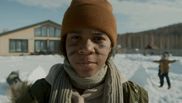 Close-up portrait shot of teenage African American girl in woolly hat and scarf standing outdoors on sunny winter day, applying war stripes on face, and children throwing snowballs in background