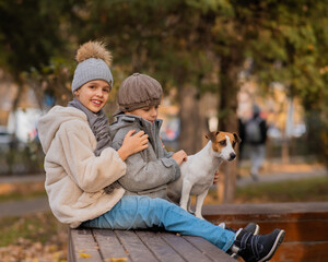 Brother and sister sit in an embrace with a dog on a bench for a walk in the autumn park. Boy, girl and jack russell terrier. 