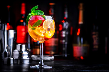 Pink sangria alcoholic cocktail drink with rose wine, peach, orange, lime and ice. Black bar counter background, steel bar tools and bottles