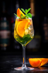 White sangria alcoholic cocktail drink with viura spanish wine, peach, orange, lime and ice. Black bar counter background, steel bar tools and bottles