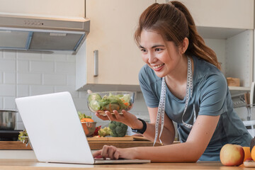 Smiling fitness nutritionist in casual clothes On the table with a laptop computer Holding a bowl of organic vegetable salad Place a measuring tape around your neck in a modern kitchen at home