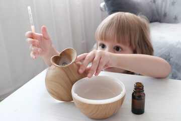 A little fair-haired girl fills a water diffuser, a wooden jug with a pipette, with aromatic oil....