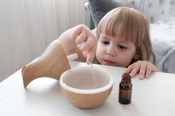 A little fair-haired girl fills a water diffuser, a wooden jug with a pipette, with aromatic oil. Concept of health care, air humidification.