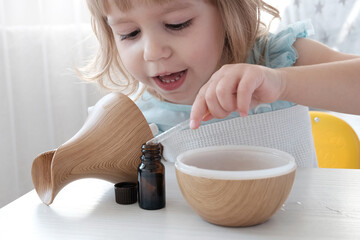 A little fair-haired girl fills a water diffuser, a wooden jug with a pipette, with aromatic oil....