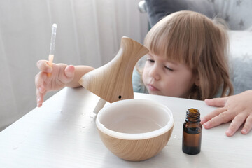 A little fair-haired girl fills a water diffuser, a wooden jug with a pipette, with aromatic oil....