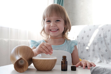 A cheerful little fair-haired girl fills a water diffuser, a wooden jug with a pipette with...
