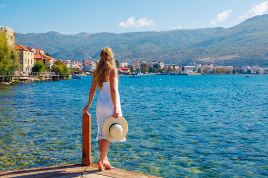 Woman in white dress standing on wooden pier looking at the sea or lake (Macedonia, Ohrid lake, Albania)