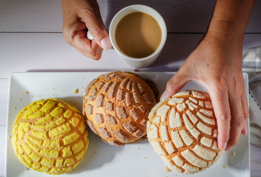 Hands of Mexican Hispanic woman . Concept of taking food with hands or handling food. Mexican concha in a hand and a cup of coffee in the other hand over a white wooden table, top view 