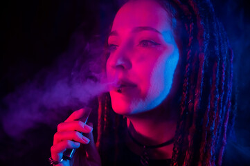Caucasian girl with dreadlocks smokes a vape in red blue light. 