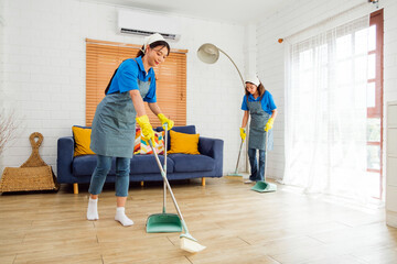 Two Asian young professional cleaning service women worker team working in the house. Girls housekeeper sweeps broomsticks on the wooden floor with another one cleaning under the sofa. Cleaner. - obrazy, fototapety, plakaty