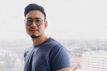 Asian man wear glasses and blue t-shirt with beard, smiling and standing over city view at condominium, looking at camera living at apartment room alone, having happy good life.