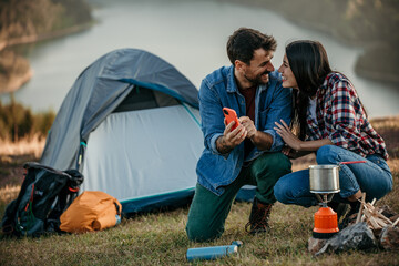 Diverse couple camping by a lake with a stove, capturing the moment on a phone