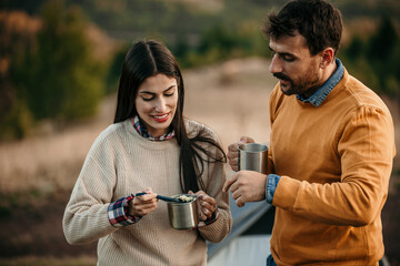 Fototapeta premium Diverse duo cooking noodles outdoors by a peaceful lake at their campsite