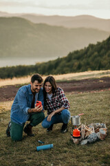 A couple using a phone, relaxes around a campfire on a hilltop, overlooking a serene lake