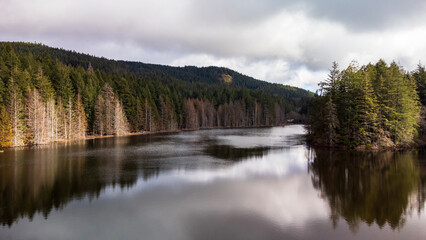 Fototapeta premium Clouds reflect off the still lake water. Forest in shadow hugs the shore. 