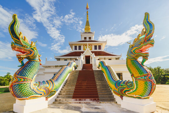 A pair of colorful Naga statues stand majestically atop the grand staircase of Wat Pha Tak Suea Temple in Sangkhom District, Nong Khai Province, Thailand, against a clear blue sky.