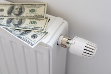 Stack of dollar banknotes on heating radiator battery with thermostat temperature regulator. Concept of expensive heating costs and rising energy bill prices for winter cold season.