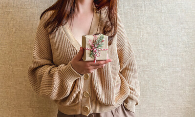 An unrecognizable brunette girl in a beige sweater holds a Christmas gift in eco packaging in her hands. a small gift in honor of Christmas, new year