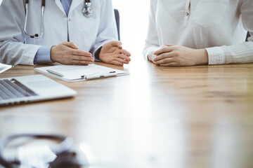 Doctor and patient discussing something while sitting near each other at the wooden desk in clinic, close up of hands. Medicine concept