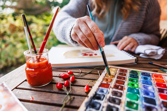Woman artist dips paintbrush into color palette and painting a watercolor illustration