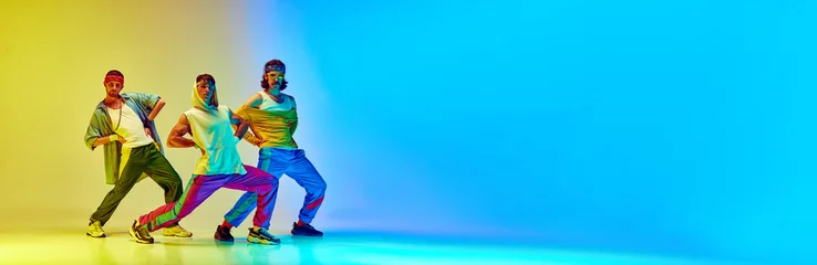 Foto op Plexiglas Hip hop dancers. Three men in stylish vintage sportswear and accessories posing against gradient yellow blue background in neon light. Concept of sportive and active lifestyle, humor, retro style. Ad © master1305