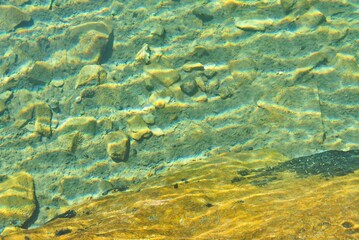Lake background texture with turquoise transparent water and light brown stones. Waves of waves