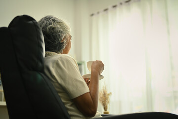 Calm and peaceful elderly woman with cup of coffee sitting on armchair and looking away