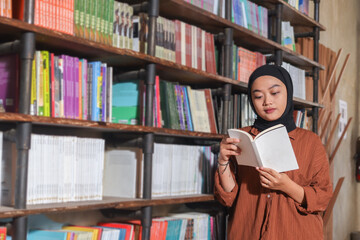 Portrait of Asian hijab woman holding book in front of library bookshelf. Muslim girl reading a...