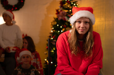 Portrait of teenage girl in Santa hat sitting on chair in front of Christmas tree, Merry Christmas and Happy Holidays!