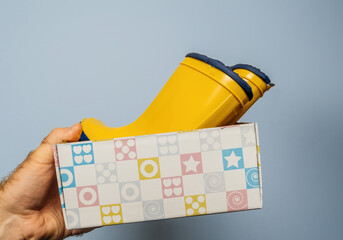 A male hand presents a meticulously packaged pair of yellow boots against a neutral gray backdrop