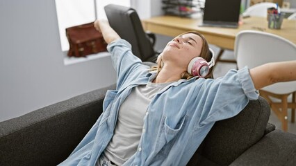Attractive confident young blonde woman enjoying work, peacefully listening to music and stretching arms in her office, showing the beautiful art of balancing business and relaxation.